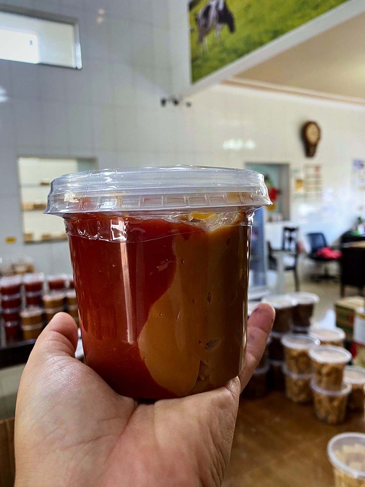Pote de doce de leite com goiabada segurado na loja, com o mural da vaquinha ao fundo